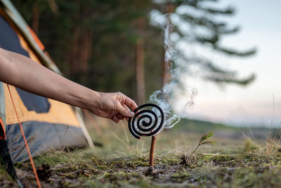 Woman installing burning mosquito repellent coil with smoke on dry tree twig in forest near tent