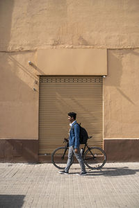 Man wheeling bicycle by wall during sunny day