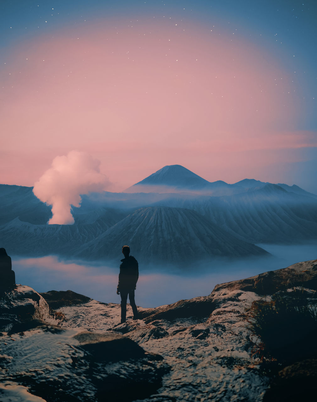 REAR VIEW OF MAN STANDING ON ROCKS AGAINST SKY