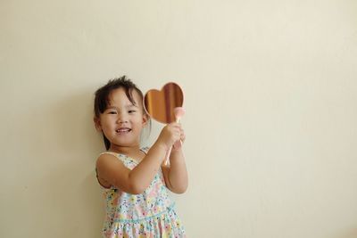 Portrait of smiling girl applying make-up while holding mirror against wall