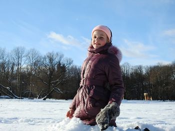 Portrait of smiling girl kneeling on snow covered field against blue sky