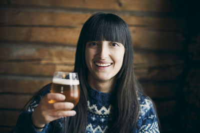 Portrait of smiling woman holding drinking beer glass while sitting at log cabin