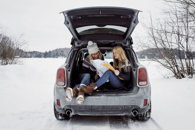 Happy female friends reading map while sitting in car trunk during winter