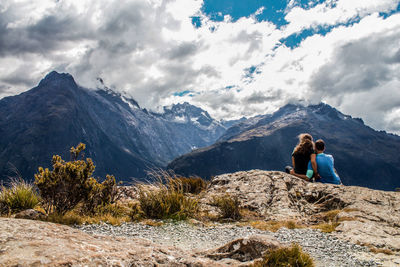 Rear view of couple sitting on mountain against cloudy sky