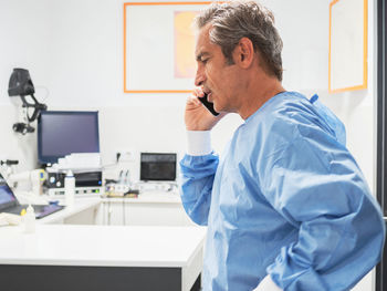 Side view of mature man in medical uniform answering phone call while working in modern hospital