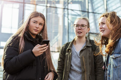 Teenage girl holding mobile phone while standing with friends against building