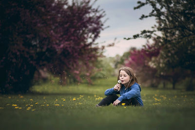 Girl blowing dandelion flower while sitting on grassy field at park