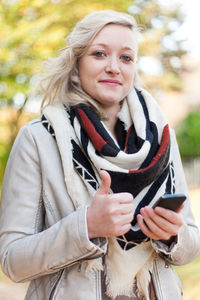 Portrait of young woman showing thumbs up while holding mobile phone