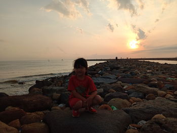 Man sitting on rock at beach against sky during sunset