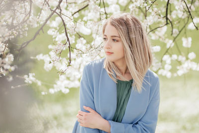 A beautiful young blonde woman standing near an apple tree in a green dress and a blue coat