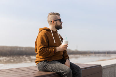 Man drinking coffee sitting on wall against sky