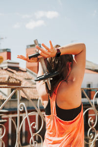 Fit woman in black and orange sportswear exercising with dumbbells on the rooftop on sunny day.