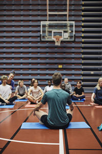 Male coach teaching yoga to male and female students in sports court