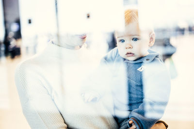 Thoughtful young mother looking trough window holding his infant baby boy child while waiting to