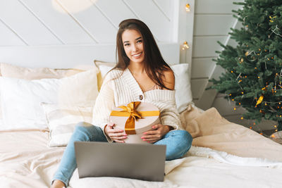 Young asian woman in white sweater with gift box on bed with laptop in room with christmas tree 
