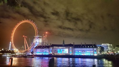 Illuminated ferris wheel by river against sky at night