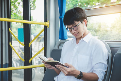Smiling young man listening to music on headphones while reading book in bus