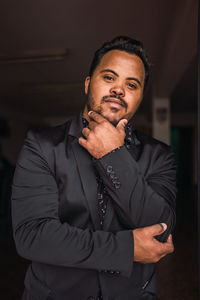 Self assured black male with beard in elegant suit standing with hand at chin and looking at camera
