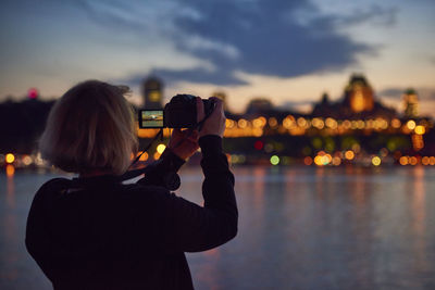 Rear view of woman photographing illuminated cityscape at sunset