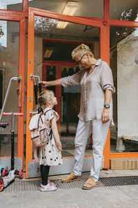 Smiling elderly woman opening door for granddaughter while standing near kindergarten