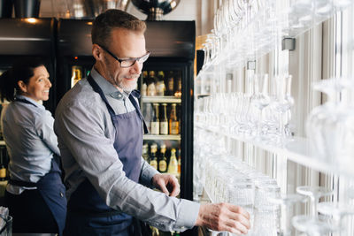 Mature man arranging wineglasses while waitress in background at restaurant