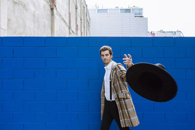 Stylish man wearing checked jacket throwing black hat against blue wall