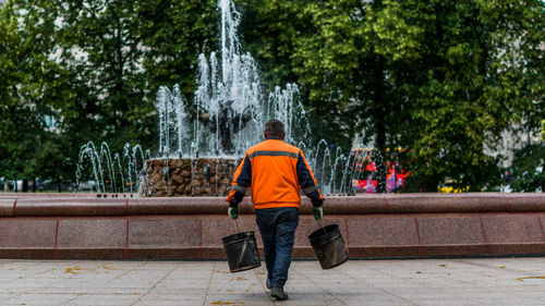 Rear view of male worker carrying buckets against fountain