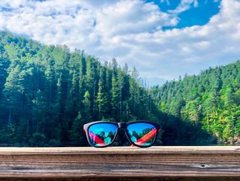 Sunglasses with blue lenses on a wooden fence with lake and green forest in the back