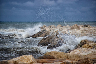 Waves splashing on rocks at sea shore against sky