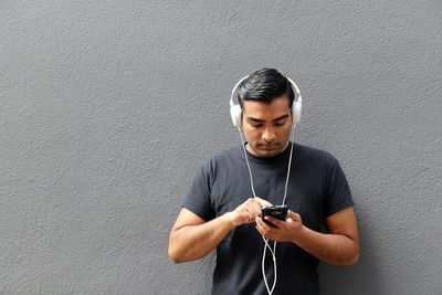 Young man using mobile phone while listening music against wall