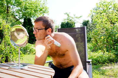 Shirtless of mid adult man shaving while looking at mirror in garden