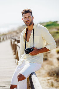 Portrait of man with camera sitting on railing against sky