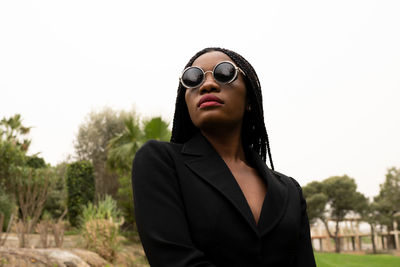 Confident african american female in stylish round sunglasses standing on street with lush green trees on summer day in city