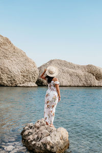 Rear view image of woman in long summer dress standing on rock on beautiful beach.