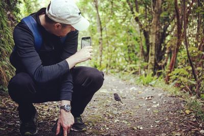 Man crouching while photographing bird on pathway in forest