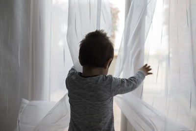 Rear view of boy standing by curtain at home