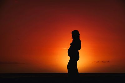 Silhouette woman standing on beach against orange sky
