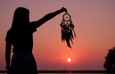 Woman holding dreamcatcher against sky during sunset