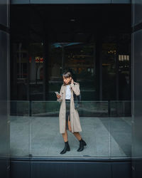 Full body of stylish female browsing cellphone while walking near contemporary building with glass fence on street of city