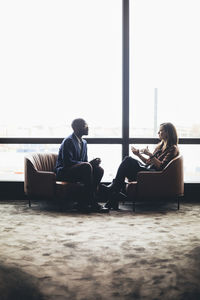 Female and male coworkers discussing while sitting against window at workplace