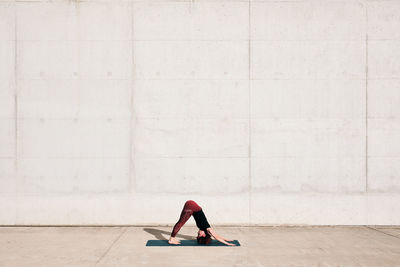 Side view of trendy fit female athlete in sportswear doing downward facing dog yoga pose on sports mat while training alone on street against concrete wall in sunny day