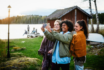 Happy female friends taking selfie with smart phone while standing on grassy field against lake during sunset