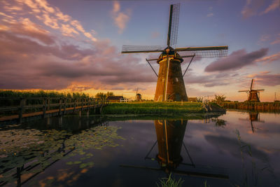 Traditional windmill by lake against sky during sunset