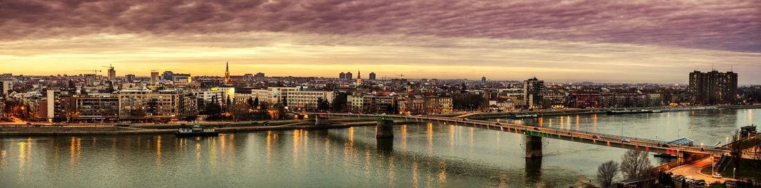 Bridge over river by buildings against sky at sunset