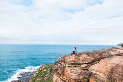 Side view of woman sitting on cliff against cloudy sky