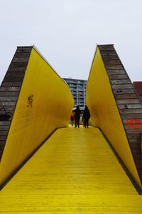 People walking on yellow bridge against sky in city