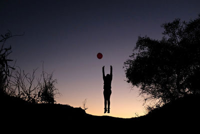 Low angle view of silhouette woman with arms raised jumping on hill against clear sky during sunset
