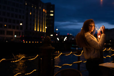Portrait of young woman holding illuminated string light at night