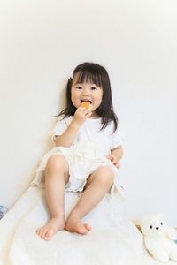 Full length of cute girl eating biscuit while sitting on bed at home