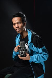 Portrait of young man holding old camera against black background